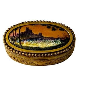 May include: Oval-shaped gold trinket box with a raised beaded edge. The lid features a painted scene of a landscape with trees, a river, and a building in the background. The colors are muted and include shades of orange, brown, green, and blue.