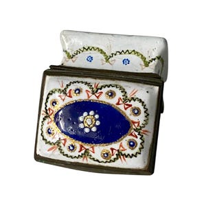 May include: A small, rectangular trinket box with a hinged lid. The box is white with a blue oval design in the center, surrounded by gold and red accents. The lid has a scalloped edge with green and blue floral details. The box has a bronze-colored metal frame.