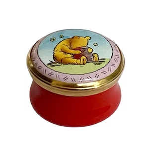 May include: A small round trinket box with a bright red base and a gold-toned metal rim. The lid features a circular illustration of a yellow bear sitting on green grass, holding a brown honey pot against a light blue sky with small bees.