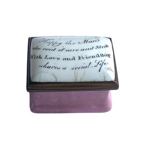 May include: A small, rectangular trinket box with a pink base and a white lid. The lid features a handwritten quote in black ink. The quote reads: "Happy the Man who void of care and Strife With Love and Friendship shares a social Life."
