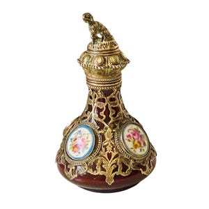 Può includere: Un flacone di profumo in vetro rosso intenso racchiuso in un ornato rivestimento in filigrana di metallo color oro. Il flacone presenta inserti circolari in porcellana con dipinti floreali e un tappo sormontato da una piccola statuetta di cane in metallo.