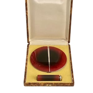 Pode incluir: Um estojo compacto vintage aberto com um compacto oval vermelho e preto e um porta-batom a condizer. O estojo é forrado com cetim creme e assenta numa superfície amarela. O estojo tem um exterior castanho padronizado.