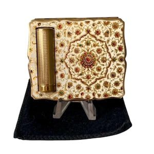 May include: A rectangular gold-tone metal compact case featuring an integrated cylindrical lipstick holder on the left side. The surface is decorated with an intricate cream-colored floral pattern accented with small red and green inlaid stones.