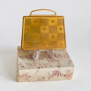 May include: A gold-toned metal compact case shaped like a handbag with a checkered pattern and small floral engravings. The compact features a top handle and rests on a rectangular cardboard box decorated with a red floral print and the text Mascot.