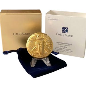 May include: A gold-toned Estée Lauder Aquarius compact featuring an embossed water bearer figure and small inset crystals. The set includes a navy blue velvet drawstring pouch, a gold outer box, and a white product box labelled Lucidity Translucent Pressed Powder, 2.8 g.