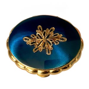 Può includere: Uno specchietto compatto rotondo con un coperchio blu profondo dalla finitura metallica, caratterizzato da un intricato ornamento in filigrana color oro al centro. Il bordo è smerlato con una finitura dorata lucida, creando una cornice decorativa.