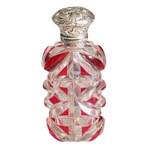 Puede incluir: Un frasco de perfume de cristal tallado en rojo y transparente con un patrón geométrico de facetas en todo el cuerpo. El frasco está rematado con una tapa redonda de metal plateado que muestra un intrincado diseño floral en relieve.