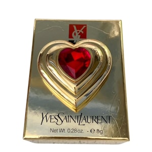 May include: A gold-toned compact with a heart-shaped design and a large, red gemstone at its center. The compact is in a rectangular box with the Yves Saint Laurent logo and text. The net weight is 0.28 oz.