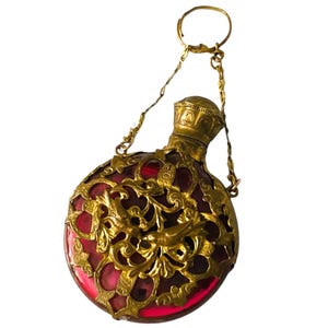 May include: A vintage perfume bottle featuring a red glass body, enclosed in elaborate gold filigree. It has a gold cap and is suspended by a fine gold chain and ring. The design is detailed, demonstrating a classic aesthetic, with a height of approximately 10 cm.