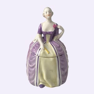 May include: A porcelain figurine of a woman in a purple and cream-colored dress, holding a pink flower and a yellow fan. The figurine is a decorative container with a lid, showcasing a vintage aesthetic.