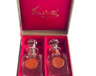 May include: A rectangular beige presentation box with a gold metal latch, containing two clear glass perfume bottles with faceted stoppers. Each bottle features a round orange wax seal with the letter G. The interior is lined in bright pink.