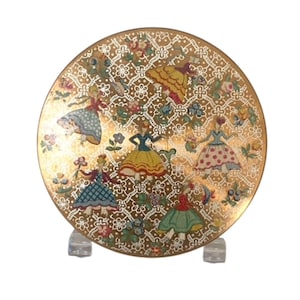 Peut inclure: Une assiette décorative ronde dorée avec un motif détaillé de danseuses en robes colorées. L'assiette présente un motif floral et dentelle, avec les personnages dans diverses poses. L'assiette est présentée sur un support transparent.