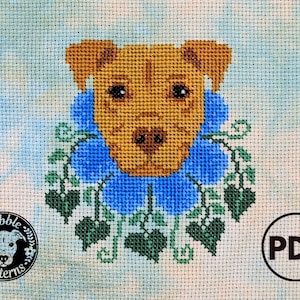 May include: Cross stitch pattern featuring a brown dog with blue flowers and green leaves around its neck. The pattern is titled "Pebble Patterns" and is available as a PDF download.
