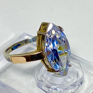 May include: A gold-toned ring featuring a large, clear, marquise-cut gemstone set in a prong setting. The band is smooth and metallic, showcasing a reflective surface that catches the light, highlighting the elongated shape of the central stone.