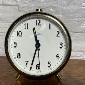 May include: A gold-toned alarm clock with black hands and black numbers on a white face. The clock reads "PRIM" and "riton".