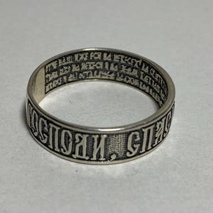 May include: A silver ring with engraved text. The ring has a band with a repeating pattern of text around the outside. The inside of the ring also has engraved text. The ring is on a white surface.
