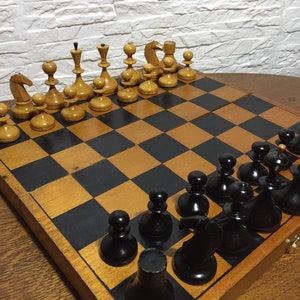 Puede incluir: Un tablero de ajedrez de madera con casillas negras y blancas. El tablero está abierto y muestra una partida en curso con piezas de ajedrez blancas y negras.