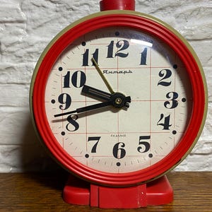 May include: A vintage red and green analog clock with a white face and black numerals. The clock face has a grid pattern and the word "Янтарь" is printed above the center. The clock has a red base and a red top.