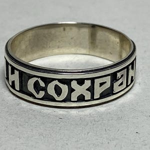 May include: A silver ring with a black band featuring Cyrillic text. The ring has a polished finish and a simple, classic design. The text is in a bold, sans-serif font.