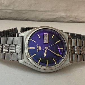 May include: A Seiko 5 wristwatch with a metallic silver-tone stainless steel link bracelet and a round case. The watch features a deep purple sunburst dial with gold-tone hour markers, gold-tone hands, and a day-date window at the three o'clock position.