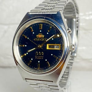 May include: A silver-tone stainless steel wristwatch with a dark blue circular dial, gold-tone hour markers, and gold-tone hands. The watch features a day-date window at the three o'clock position, the Orient logo, and the text Crystal 21 Jewels.