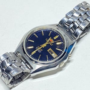 May include: A silver-toned wristwatch with a dark blue face and gold-colored hands and markers. The watch has a date display showing "MON 31". The watchband is made of silver-toned metal links.