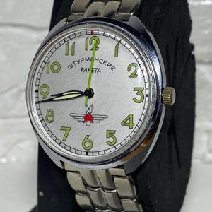 Peut inclure: Une montre-bracelet argentée avec un cadran blanc et des aiguilles noires. La montre a un bracelet en métal argenté et les mots "Штурманские Ракета" imprimés sur le cadran.