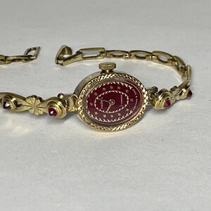 Può includere: Un orologio da polso color oro con quadrante rosso e bracciale a maglie. Il quadrante dell'orologio ha un motivo a pois ed è circondato da una lunetta dorata. Piccole gemme rosse sono incastonate nel bracciale.