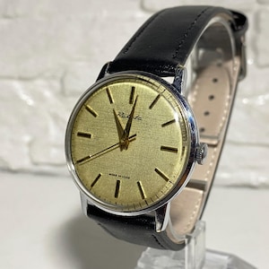 Może przedstawiać: Zegarek na rękę Raketa w stylu vintage ze złotą tarczą i czarnym skórzanym paskiem. Zegarek ma srebrną kopertę i teksturowaną tarczę z czarnymi znacznikami godzin. Na tarczy widnieje napis "MADE IN USSR".