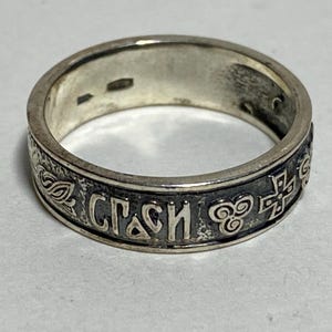 May include: A silver ring with engraved Cyrillic text and decorative patterns. The ring features a band with detailed carvings, including a cross and other religious symbols. The ring is a piece of jewellery.