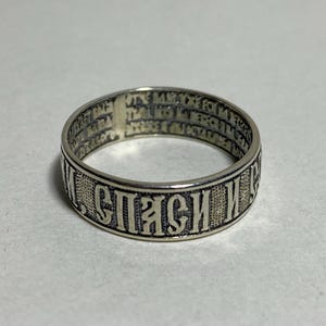 May include: A silver ring with engraved text. The ring has a band with a repeating pattern of text around the outside. The ring is circular and appears to be made of metal.