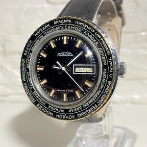 Puede incluir: Reloj de pulsera Takema vintage con esfera negra, detalles dorados y correa de cuero negro. El reloj tiene un bisel giratorio con nombres de ciudades. La fecha se muestra a las 3 en punto.