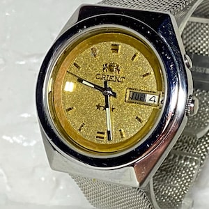 May include: A silver-toned Orient watch with a gold-colored face and a mesh band. The watch face displays the Orient logo, the day of the week, and the date. The watch has a stainless steel back.