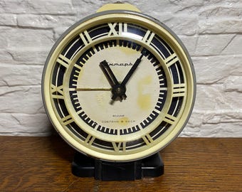 May include: Vintage analog clock with a cream-colored face, black Roman numerals, and black hands. The clock has a black base and a light yellow frame. The face has text in a foreign language. The clock is on a wooden surface.