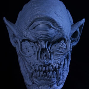 May include: A blue, detailed, sculpted mask of a creature with a single, large eye, pointed ears, and a fanged mouth.