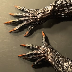 May include: Two black, clawed hands with orange tips on the nails. The hands are posed as if reaching out.