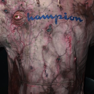 May include: A pink Champion hoodie with a large eye graphic and several holes throughout. The hoodie is stained with a dark brown and red pattern.