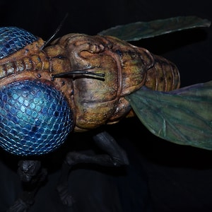 May include: A large, detailed, sculpted insect with a brown body, large blue eyes, and green wings. The insect is posed in a flying position.