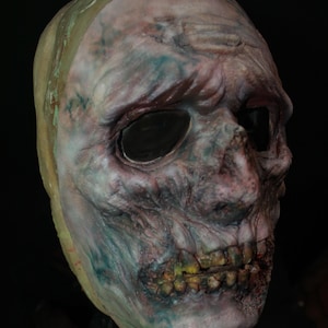 May include: A full-head mask featuring a decaying, zombie-like appearance with textured, pale skin tones, deep-set dark eye sockets, and exposed, discolored teeth. The surface displays mottled blue and red undertones with a weathered, worn finish.