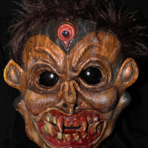 May include: A grotesque mask with a demonic appearance. The mask features a weathered, textured surface with shades of brown, orange, and gold. It has large, black eyes, sharp teeth, and a third eye on the forehead. Dark brown faux hair is attached to the top of the mask.