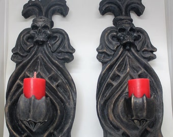 May include: Two black metal wall sconces with ornate designs, each featuring a skull and a candle holder. The sconces are decorated with intricate details and have a gothic aesthetic.