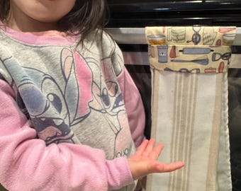 May include: A young girl in a pink and grey sweatshirt with a cartoon character design is standing in front of a stainless steel oven. A white and beige striped kitchen towel is hanging from the oven door with a colorful fabric pocket on top. The pocket has a design of various kitchen tools and utensils.