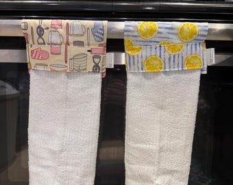 May include: Two white kitchen towels hanging on a silver rack. The towel on the left has a yellow and pink patterned fabric with images of kitchen tools and utensils. The towel on the right has a blue and yellow patterned fabric with images of lemons.