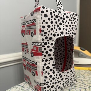 May include: A fabric storage bag with a handle. One side features a pattern of red fire trucks on a white background. The other side and handle have a black and white dalmatian spot pattern. The bag has a rectangular opening.