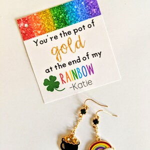 Könnte beinhalten: Eine Karte mit einem Regenbogen-Glitzerhintergrund und dem Text "You're the pot of gold at the end of my rainbow -Katie". Ein Paar goldene Ohrringe mit einem Kleeblatt-Anhänger, einem Topf-Gold-Anhänger und einem Regenbogen-Anhänger.