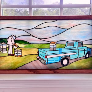May include: Stained glass artwork depicting a beekeeper tending to beehives next to a light blue pickup truck. The scene is set in a field with a mountain range in the background, framed in a brown wooden frame.