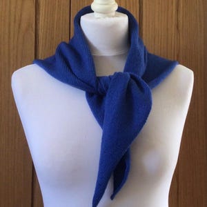 May include: A vibrant royal blue scarf, knotted at the front, is displayed on a white mannequin. The scarf's ends hang down, and it appears to be knitted. The background features a wooden wall, providing a neutral backdrop.