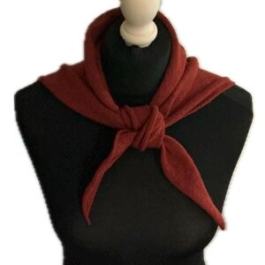 May include: A solid rust-coloured triangular scarf made of a soft, textured knit fabric. The scarf is tied in a simple knot at the centre front, creating two pointed ends that drape downwards. The fabric has a visible, slightly fuzzy knit texture.