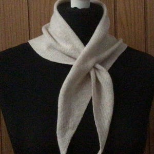 May include: A light beige scarf is draped around a black form. The scarf is knotted at the front, with the ends hanging down. The scarf appears to be made of a soft, knitted material. The background is a wooden wall.