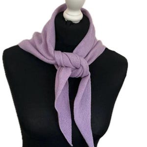 May include: A light purple knitted scarf tied in a simple knot around the neck. The scarf features a fine, textured knit pattern and pointed ends, displaying a soft, solid lavender hue that drapes evenly across the chest area.